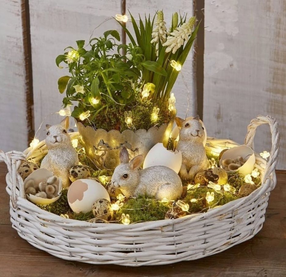 Arranjo para decoração de páscoa em cesto de vime branco com luzes de led, coelhinhos de brinquedo e cascas de ovos brancos.