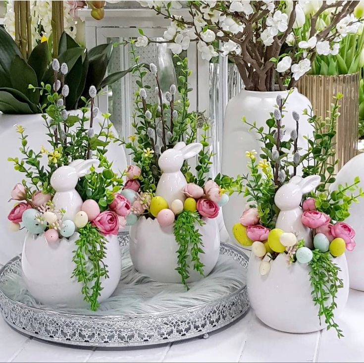 3 vasos iguais de porcelana branca com coelho de porcelana branca enfeitados com pequenos ovos colorifos, mini rosas e galhos secos.