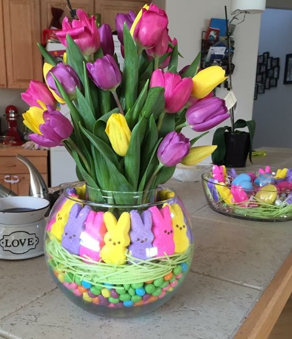 arranjo de páscoa colorido com flores, coelhinhos, fitas de papel crepom e pastilhas de chocolate em um vaso de vidro.